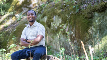 Julius Mbatia istuu metsässä kannon päällä, taustalla kivi.