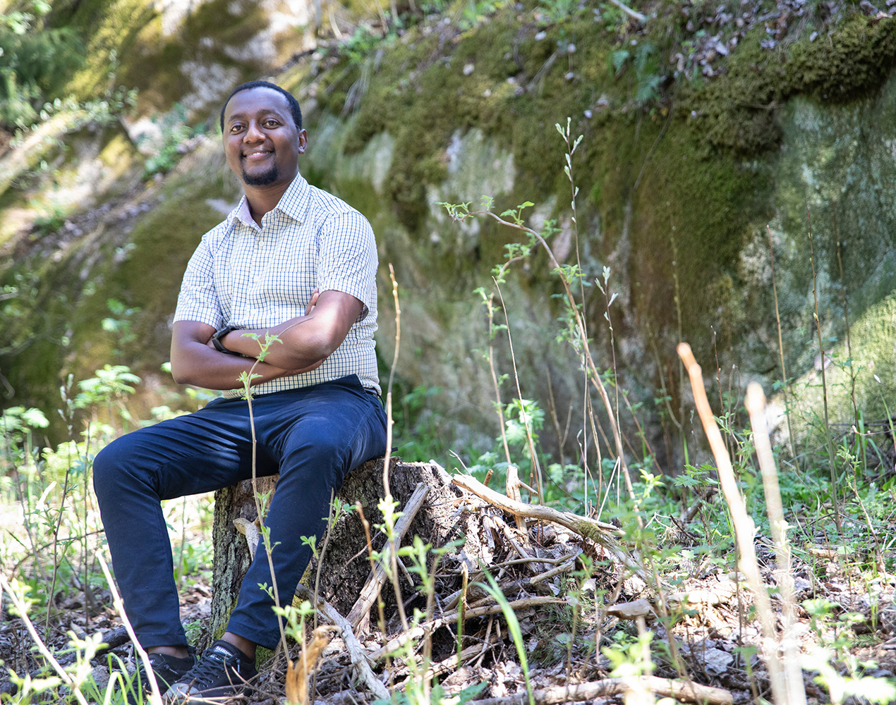 Julius Mbatia istuu metsässä kannon päällä, taustalla kivi.