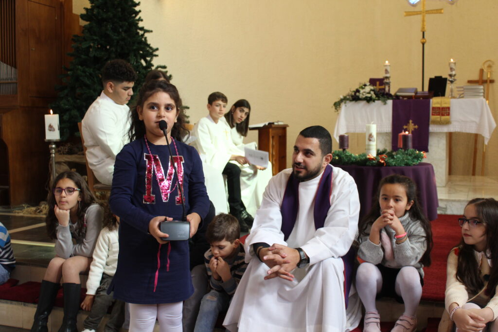 Pappi istuu kirkon etuosassa portailla lasten kanssa. Yksi lapsista puhuu mikrofoniin. Taustalla näkyy nuoria albat päällä.