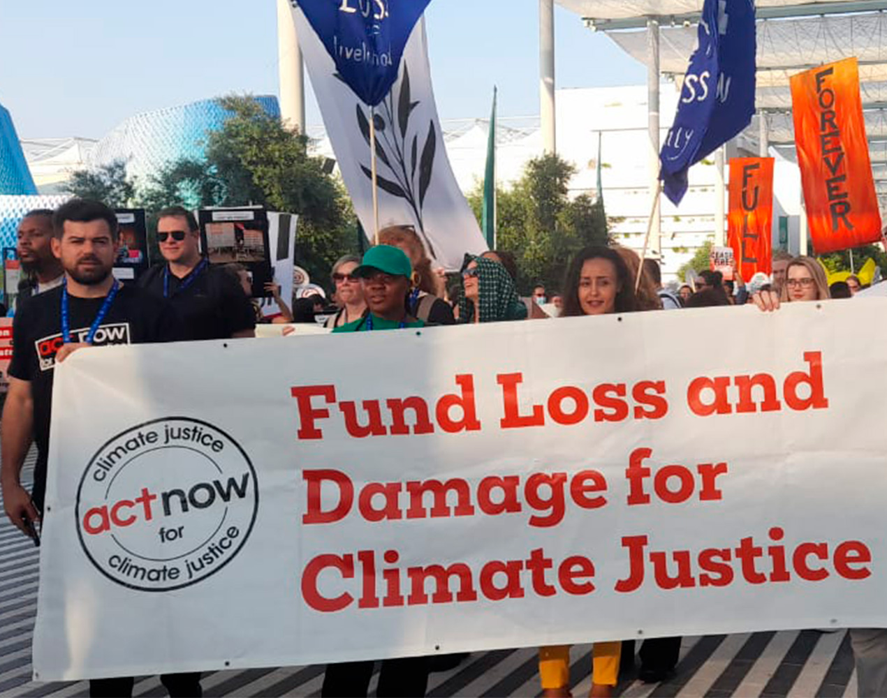 ihmisiä marssimassa ulkona mielenosoituskylttien kanssa. Isossa kyltissä lukee Fund Loss and Damage for Climate Justice.