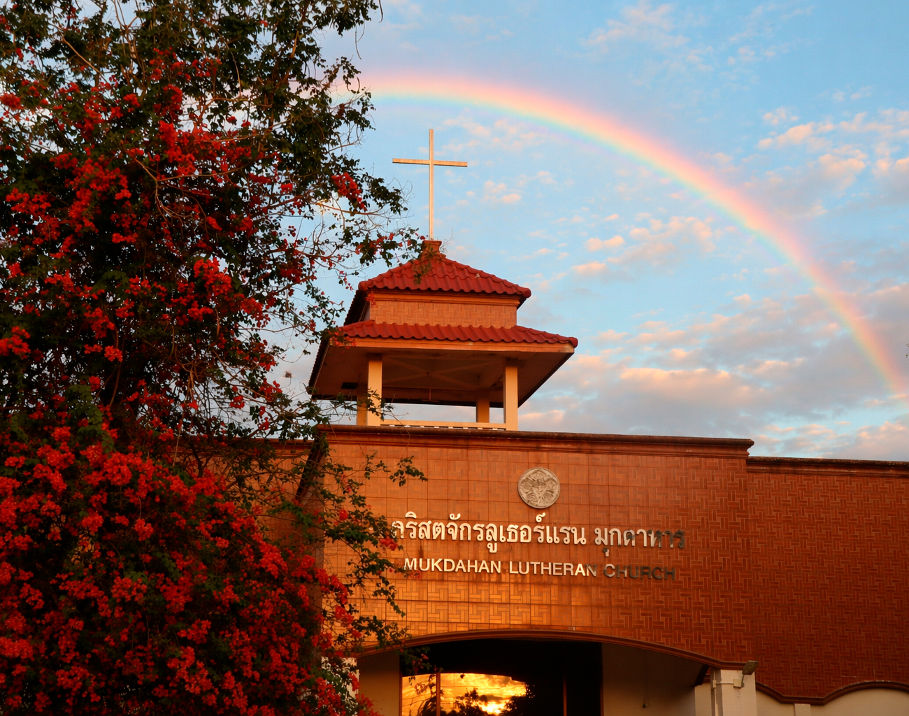 Kirkkorakennus ilta-auringossa, seinällä lukee "Mukdahan Lutheran Church". Kirkon takana taivaalla näkyy sateenkaari.