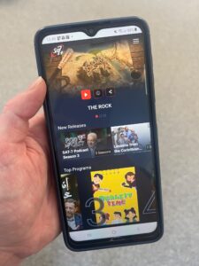 Kuvassa näkyy puhelin ja käsi. Puhelimen näytöllä näkyy tv-ohjelmien kuvia.