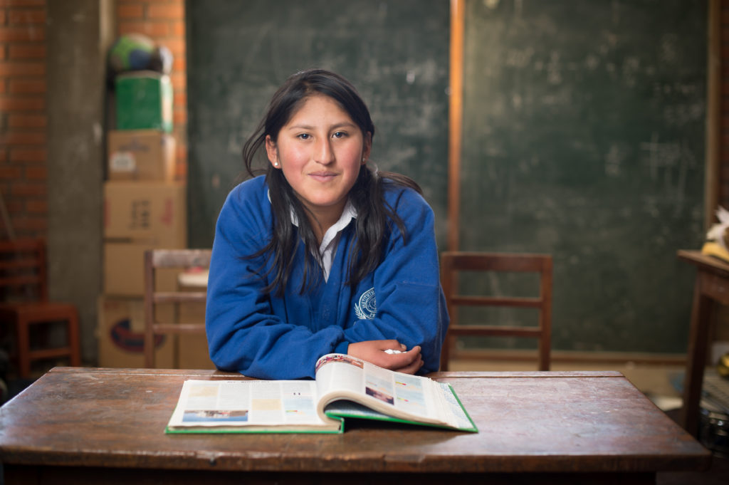 Bolivialainen koulutyttö poseeraa oppikirjan ja pulpetin ääressä koulupuvussaan.