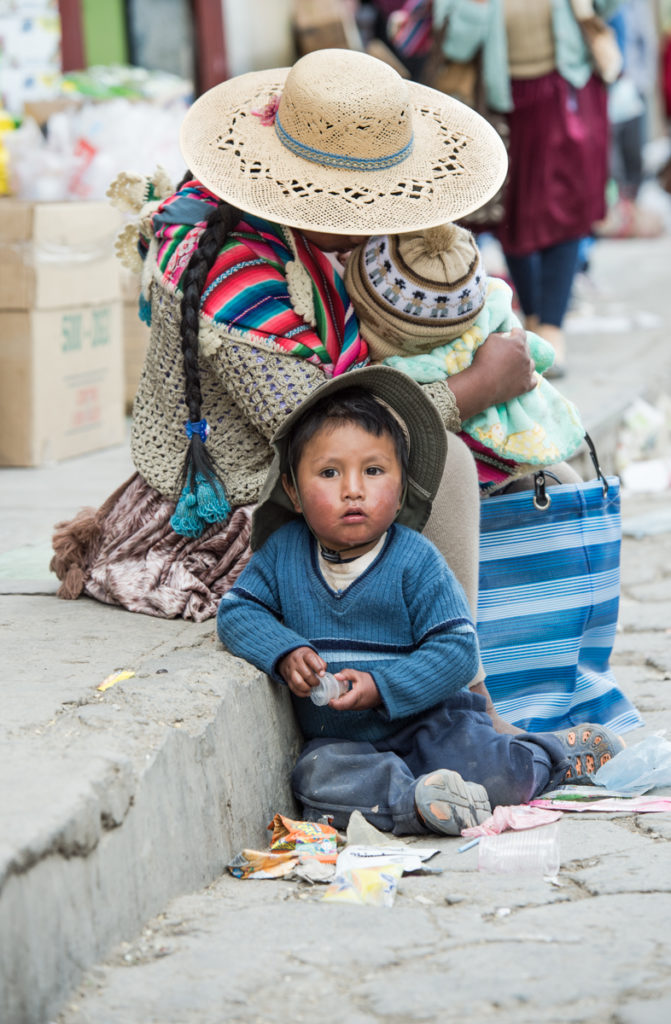 Sombreromaisen hatun alle piiloutunut nainen istuu kadun reunalla ja pitelee sylissään pientä lasta. Toinen pieni lapsi katsoo kameraan naisen vieressä.