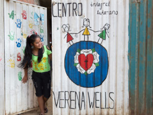 Nuori tyttö kurkistaa Verena Wells -oppilaskodin portista. Porttiin on maalattu värikkäitä kuvia ja oppilaskodin nimi.