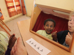 Peili, josta heijastuu iloinen etiopialainen poika. Peilin edessä on lappu, jossa on kirjoitusmerkkejä.