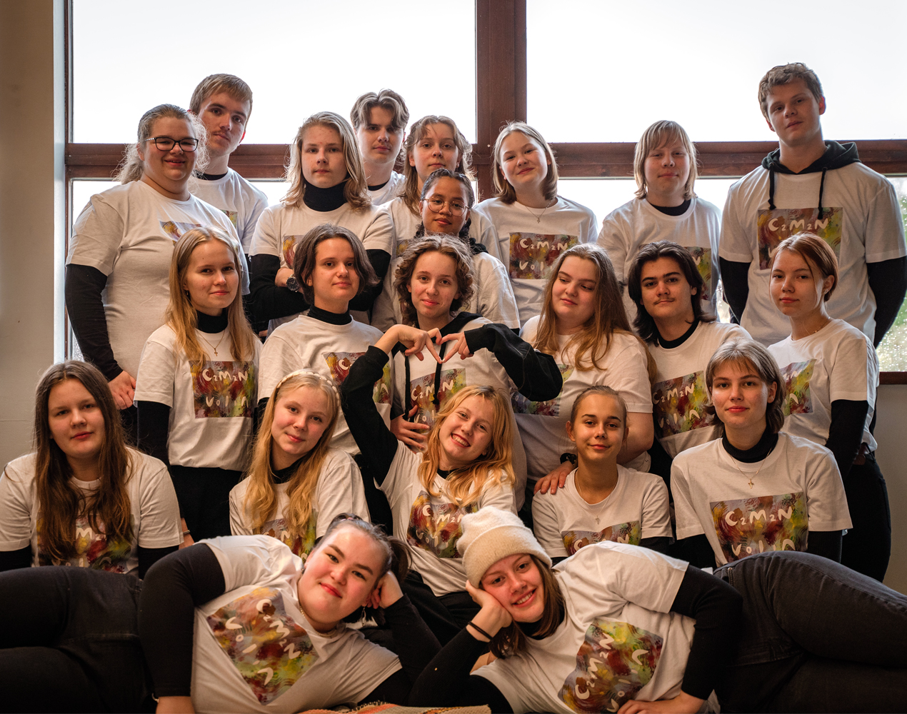 Parikymmentä parikymppistä iloista nuorta ryhmäkuvaan pakkautuneena valkoiset musiikkiryhmän t-paidat päällään.