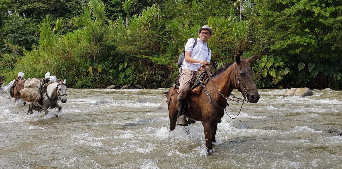 Tummahiuksinen mies ratsaasta vuolaasti virtaavan joen poikki hevosella.