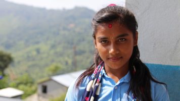 Koulupukuinen Manisha katsoo kameraan hieman hymyillen. Taustalla näkyy vehreitä kukkuloita ja rinteessä oleva rakennus.