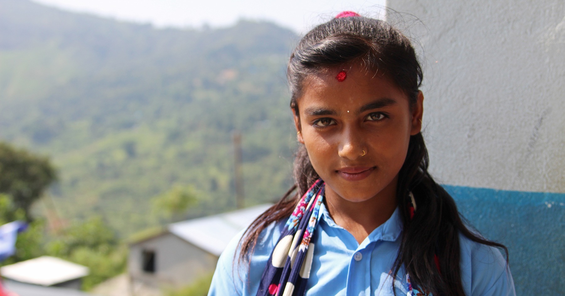 Koulupukuinen Manisha katsoo kameraan hieman hymyillen. Taustalla näkyy vehreitä kukkuloita ja rinteessä oleva rakennus.