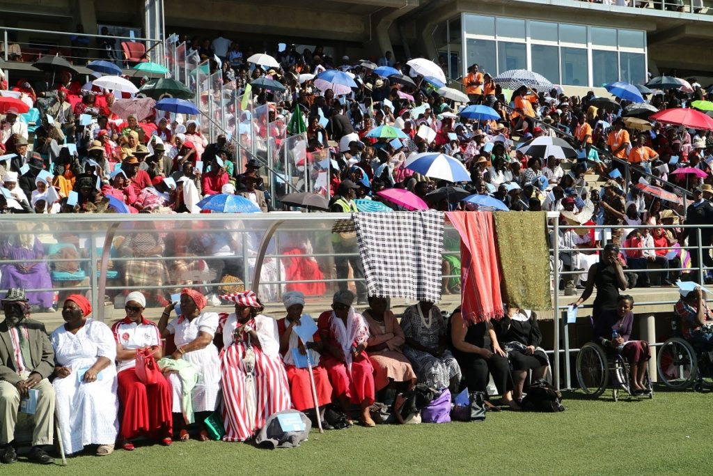 Värikkäästi pukeutunutta kansaa päivänvarjoineen stadionin katsomossa.