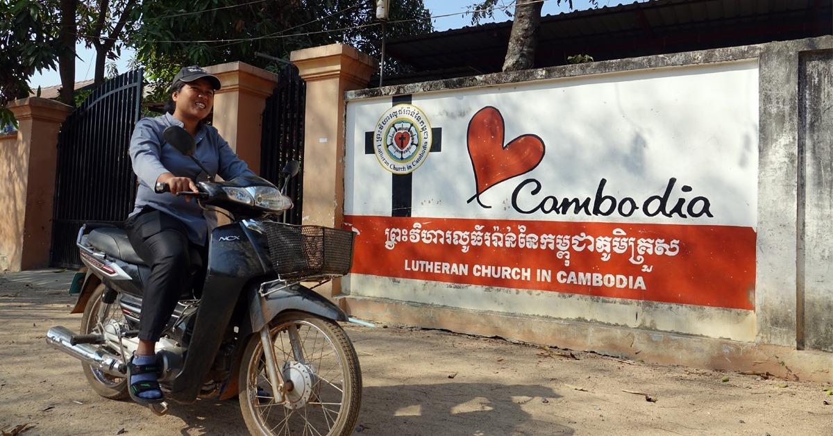 Nainen ajaa mopolla muurin viertä. Muurissa lukee Kambodzan luterilainen kirkko ja on maalattuna risti ja sydän.