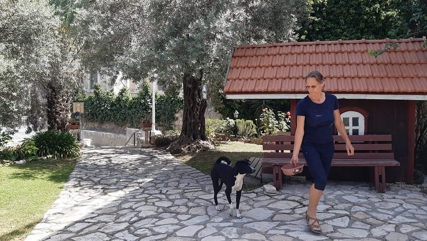 Kuvassa mustaan T-paitaan ja shortseihin pukeutunut nainen kävelee ulkona keskuksen pihalla koirankuppi kädessä, koira seuraa. Aurinko paistaa.