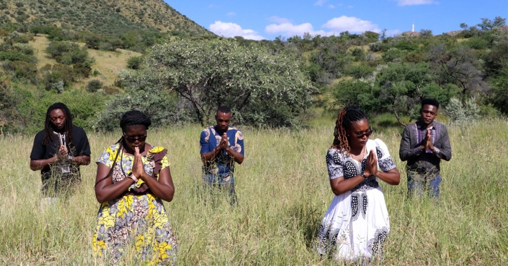Viisi ihmistä kädet rukoukseen liittyneinä Namibiassa. He seisovat pitkässä ruohikossa, taustalla metsää ja vuoren rinne.
