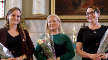 Kolme naista katsoo kirkossa hymyilleen kameraa, ruusut käsissä.
