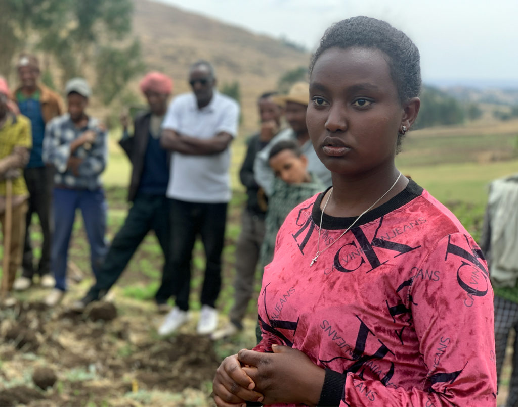 Punapaitainen etiopialaisnainen seisoo etualalla, taustalla miehiä pellonlaidalla.