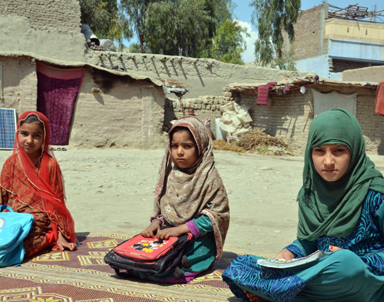 Kolme afganistalaistyttöä istuu maassa koullureput ja -kirja sylissään.