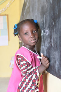 Senegalilainen pikkutyttö nojaa poskeaan liitutauluun ja katsoo kohti kameraa. Käsessä on liitu ja päällä pinkki liivi, esikoulupuku.