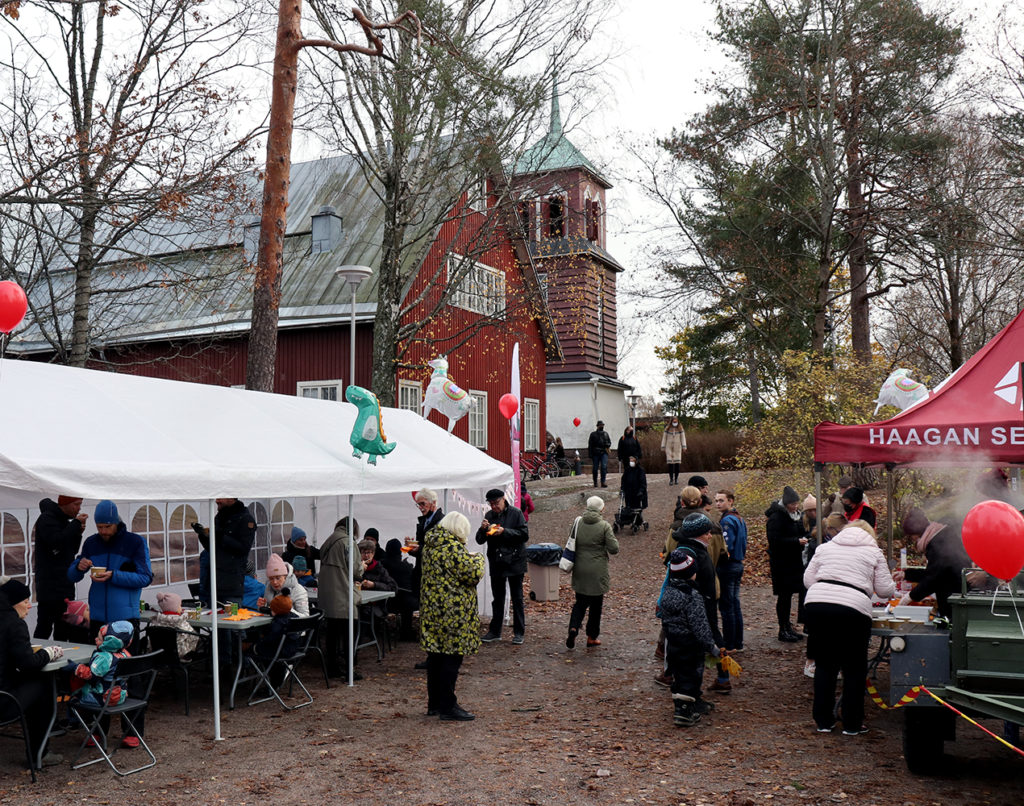 Kuva kirkon pihalta, takana punainen puukirkko, soppatykki ja telttakatos, jossa lapsiperheitä ruokailemassa.