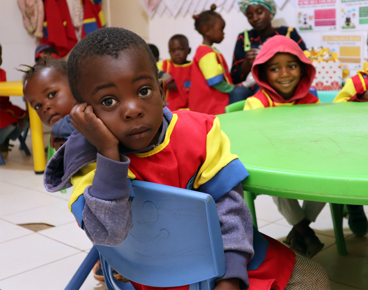 Angolalaisia päiväkotilapsia värikkäissä asuissaan pyöreän pöydän ympärillä. Etualalla pikkupoika nojaa kättään poskeensa ja katsoo suoraan kameraan.