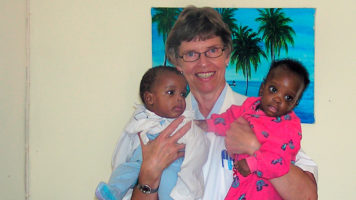 Valkotakkinen nainen pitelee hymyillen kahta lasta sylissään.