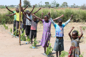 Tansanialaislapsia jonossa pituusjärjestyksessä kuvin oman puuntaimensa vieressä kädet ylhäällä voiton merkkinä.