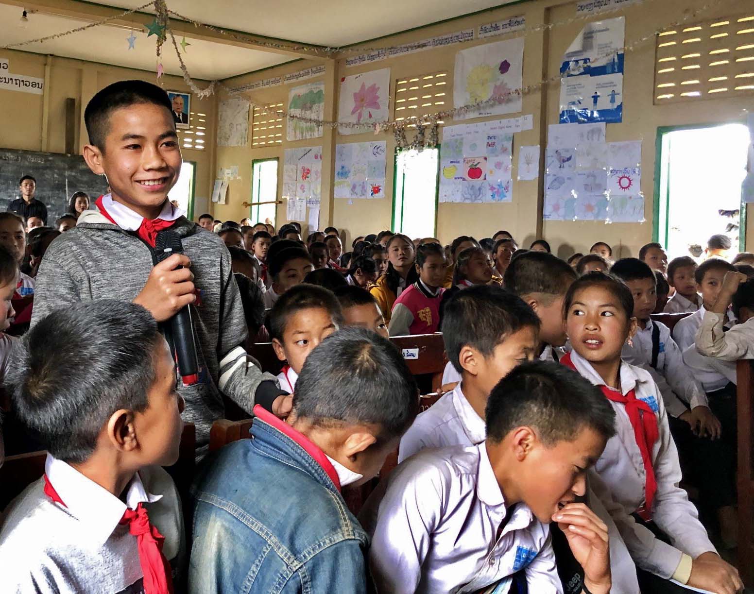 Luokkahuoneessa lapset istuvat pulpettien ääressä. Yksi lapsista seisoo mikrofoni kädessä hymyillen.