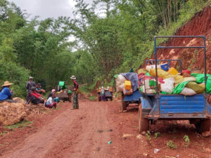 Ihmisiä ja täyteen lastattuja pieniä kuorma-autoja hiekkatien reunoilla Myanmarissa.