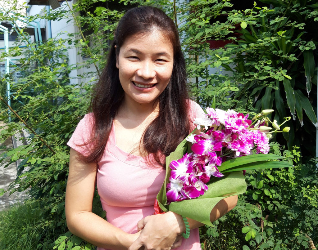 Thaimaalainen nainen pinkissä teepaidassa kukkakimppy sylissään.