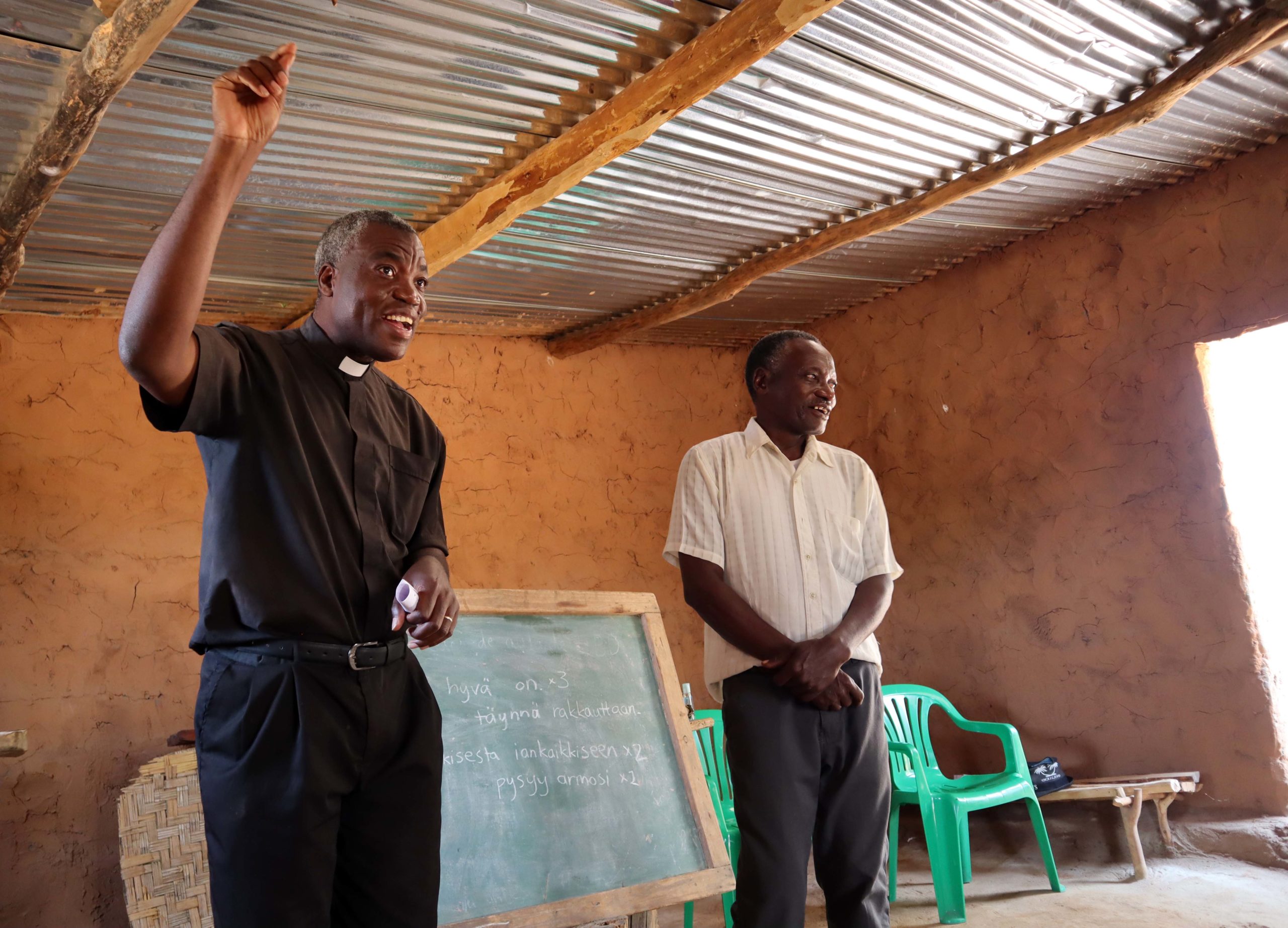 Innostunut pastori saarnaa käsi pystyssä yksinkertaisessa peltikattoisessa kirkossa, toinen seisoo vieressä tulkkaamassa.