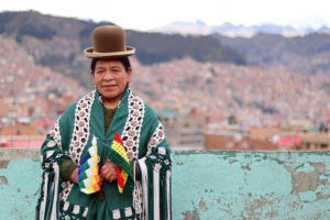 Knallihattuun ja värikkäisiin vaatteisiin pukeutunut bolivialaisnainen taustanaan vuore nrinteitä reunustavat punatiiliset talot.