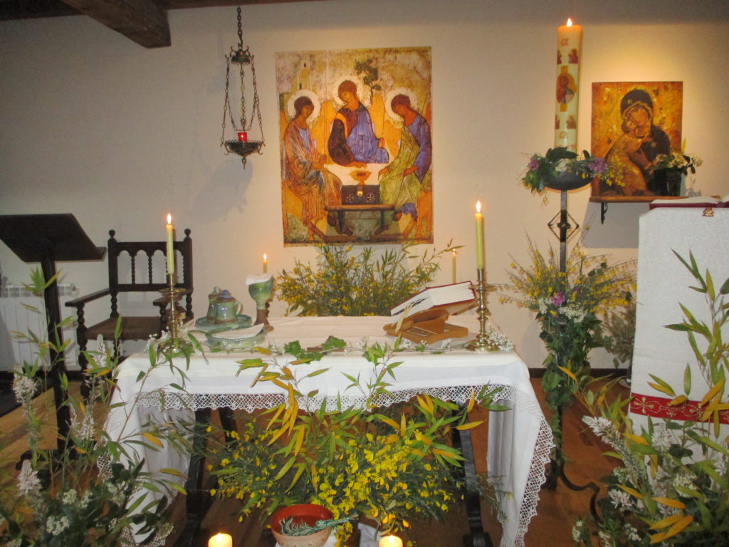 Kuvassa pöytä, jolla valkoinen liina, kynttilöitä, kasveja, takana seinällä iso ikonit.