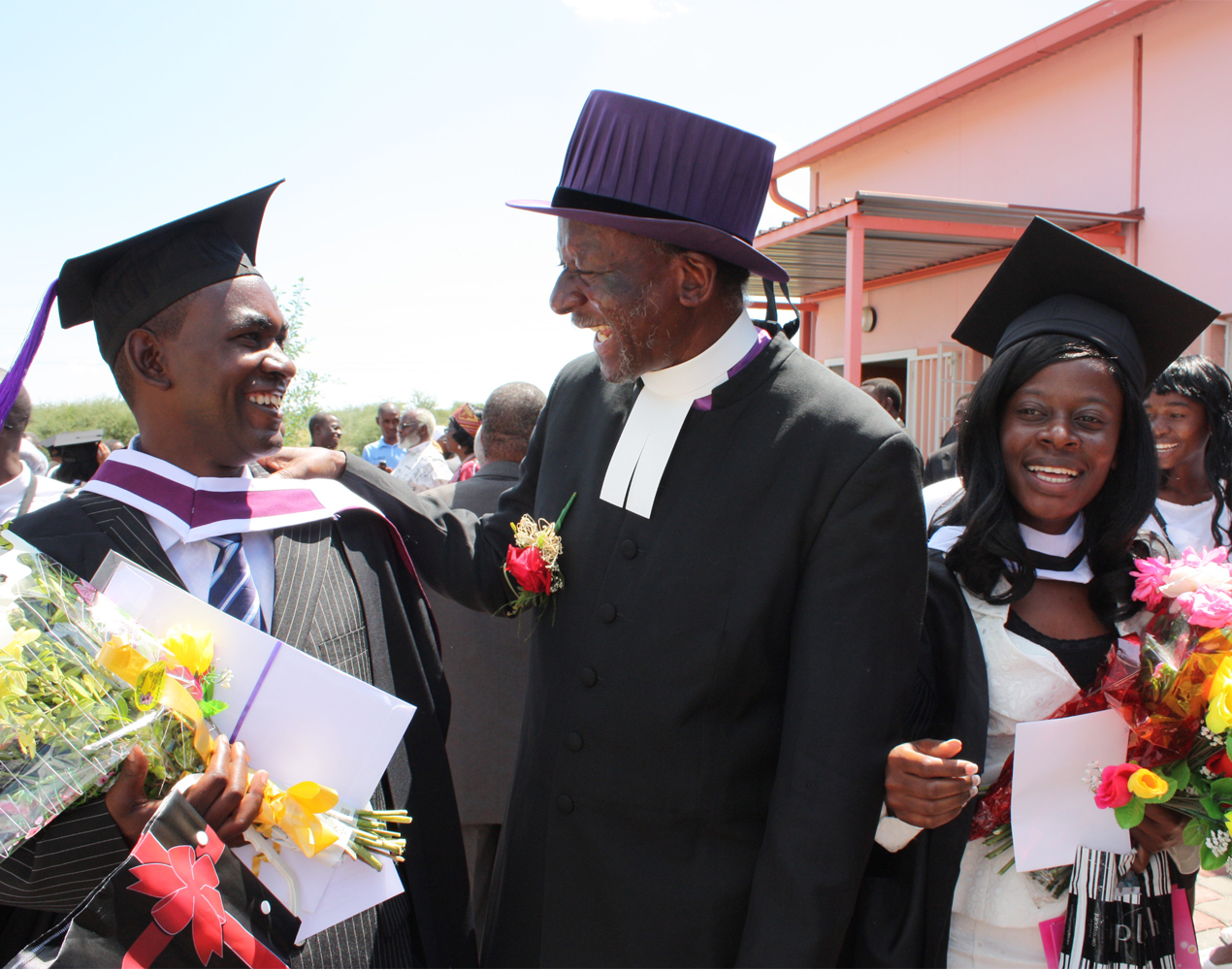Keskellä naurava papinasuun ja tohtorinhattuun pukeutunut Namibian evankelis-luterilaisen kirkon piispa molemmilla puolillaan iloiset vastavalmistuneet teologit.