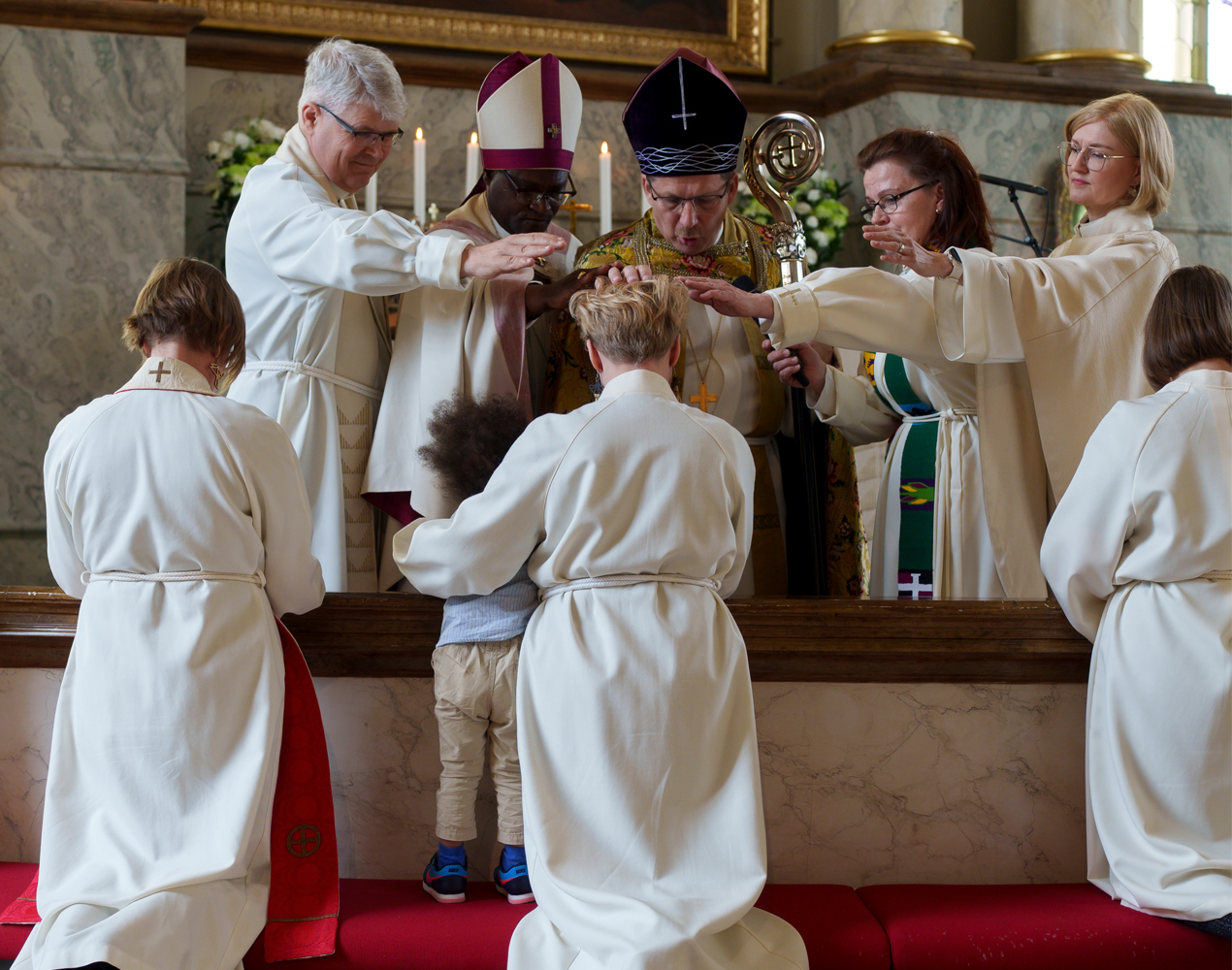 Albapukuisia ihmisiä polvistuneena alttarikaiteelle ja kaksi piispaa ja kolme muuta ihmistä siunaamassa heitä .