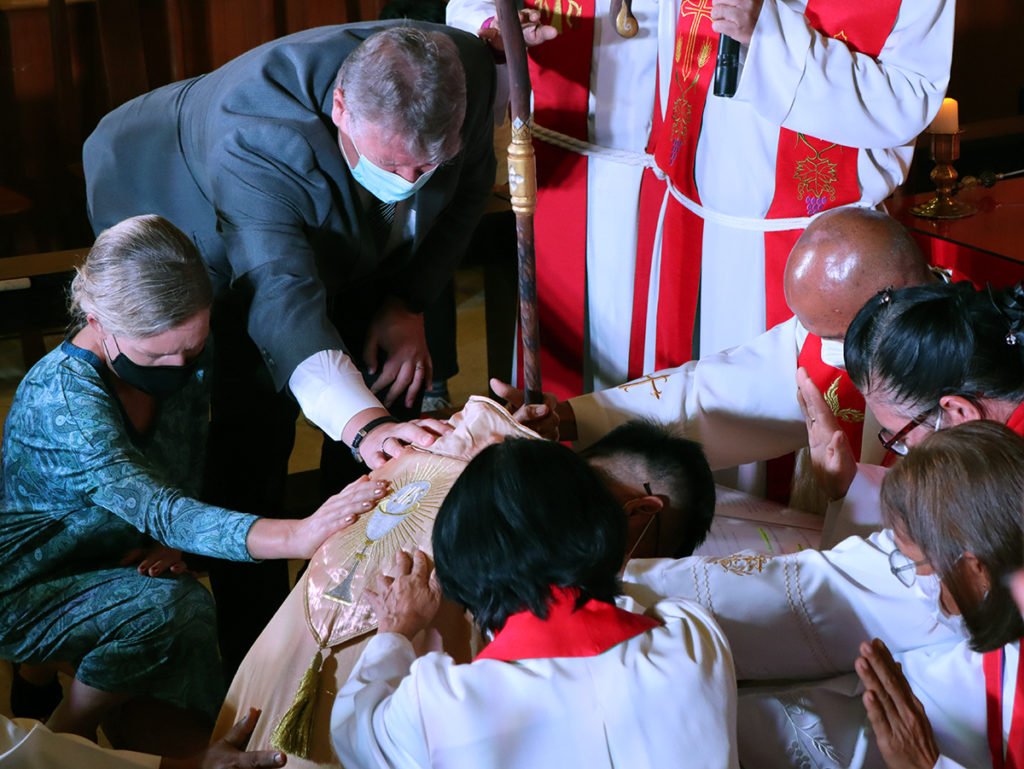 Olli Pitkänen ja muita ihmisiä ovat asettaneet kätensä kumartuneen piispan ylle.