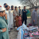 Miehiä ja lapsia on kerääntynyt katsomaan avustustiimin tuomia tarvikkeita. Kuvassa näkyy riisisäkkejä ja peittoja.