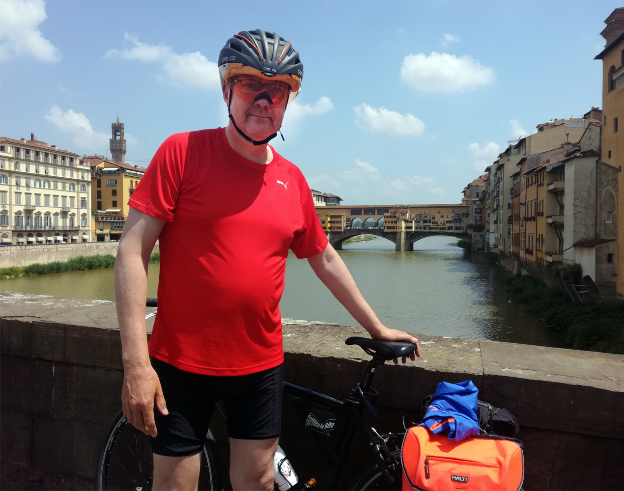 Pyöräilykypärään, punaiseen teepaitaan ja musiin shortseihin pukeutun mies nojaa pyöräänsä taustalla Firenzen kuuluisa silta.