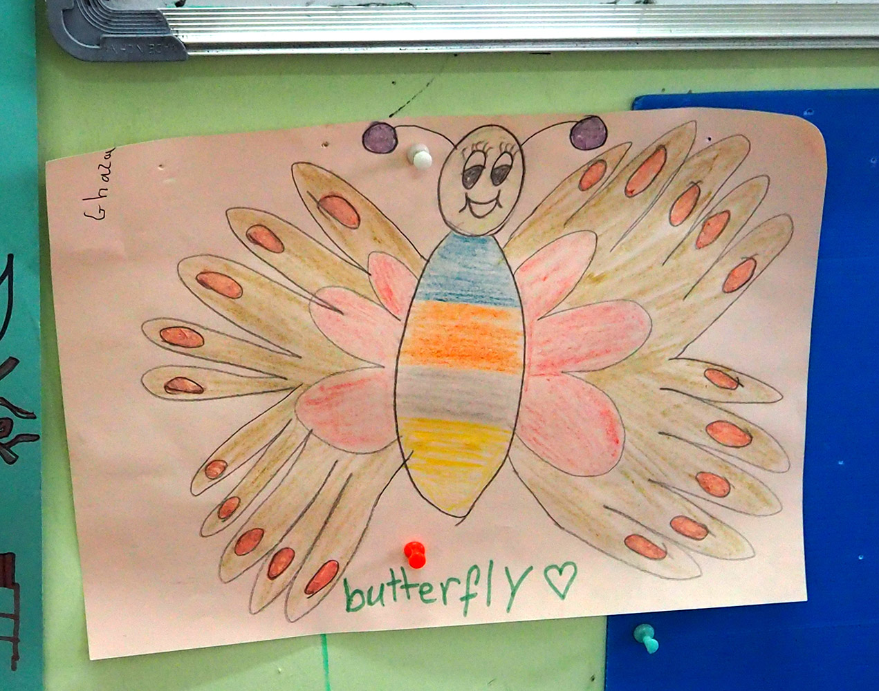 Kuvassa on piirretty perhonen, jolla on iloiset kasvot, raidallinen keskivartalo ja punaruskeat siivet joiden kärjissä on oranssit pallot.