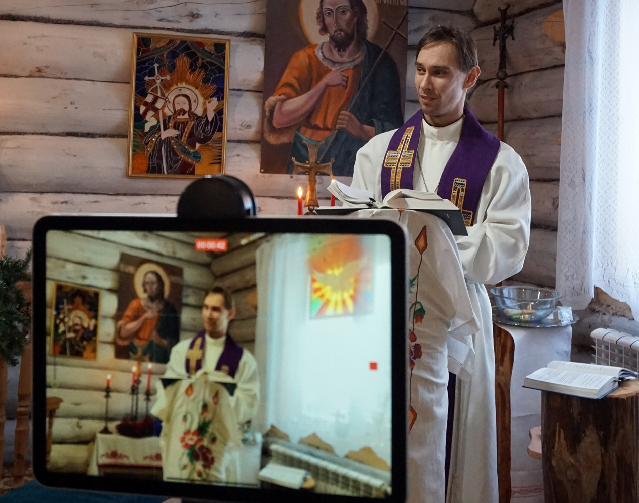 Albaan ja stolaan pukeutunut pappi puhuu jumalanpalveluksessa, taustalla hirsiseinää, papi edessä striimauslaitteet