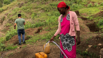 Mäenrinteellä seisova etiopialaisnainen laskee vettä letkulla keltaisiin kanistereihin