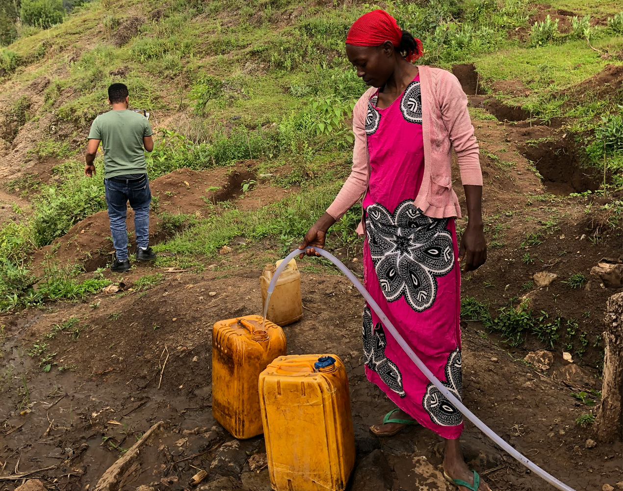 Mäenrinteellä seisova etiopialaisnainen laskee vettä letkulla keltaisiin kanistereihin