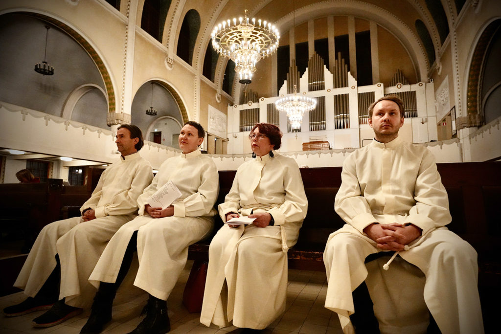 Neljä alpoihin pukeutunutta ihmistä istuu kirkon penkissä.