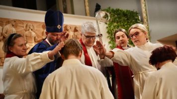 Piispa ja neljä muuta ihmistä siunaavat alttarilla albaan pukeutunutta nuorta miestä.