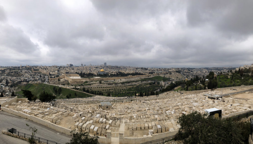 Kuvan ottaja on mäen päällä. Alempana näkyy vaaleita hautoja, sitten vihreää laaksoa ja kauempana valtavasti rakennuksia eri ajoilta: Jerusalemin vanhakaupunki vaalean muurin kera ja kauempana korkeita moderneja taloja. Pilvet peittävät taivaan.