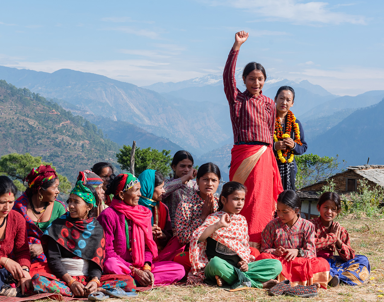 Ryhmä nuoria istuu maassa, yksi nuori seisoo ja on nostanut kätensä pystyyn. Taustalla näkyy Nepalin lumihuippuisia vuoria.