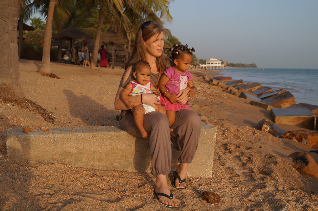 Nainen istuu rannalla kaksi pientä tyttöä sylissään. Kuvassa näkyy myös palmuja, hiekkaa ja vettä.