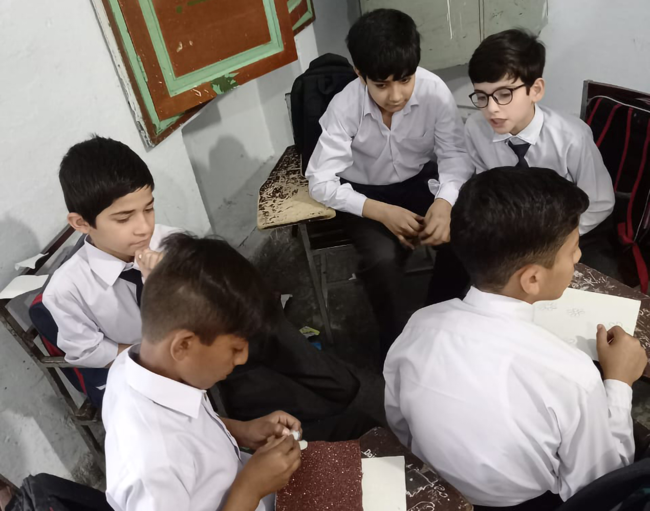 Viisi poikaa valkoisissa kauluspaidoissa. Pojat istuvat koulupöytiensä ääressä.