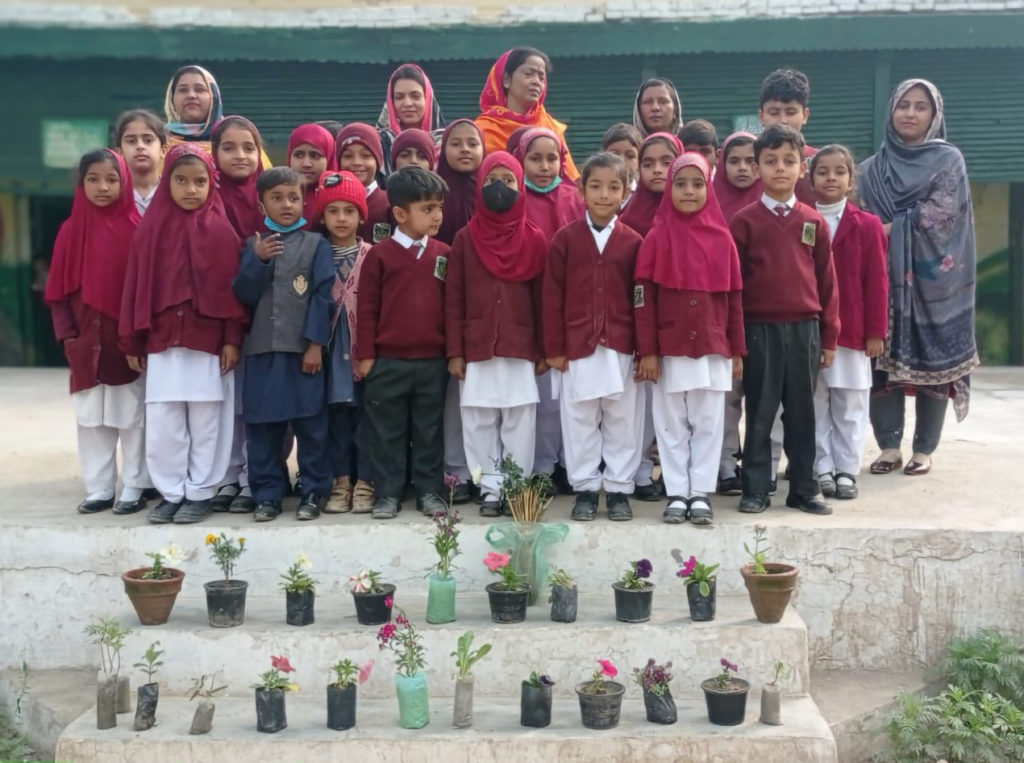 Ryhmä pieniä koululaisia ja muutama aikuinen seisovat kuvassa. Koululaisilla on punaiset koulupuserot. Pojilla on mustat housut ja tytöillä valkoiset. Lasten edessä on kukan taimia.