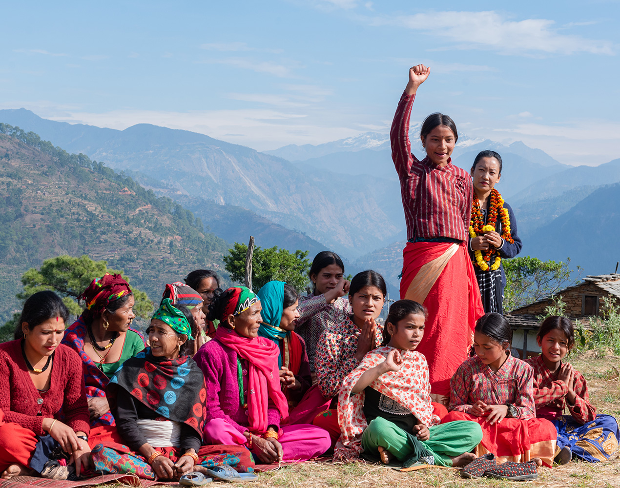 ryhmä nepalilaisia nuoria istuu maassa, kaksi heistä seisoo ja toisella seisovista nuorista on toinen käsi nostettu pystyyn, taustalla näkyy korkeita vuoria