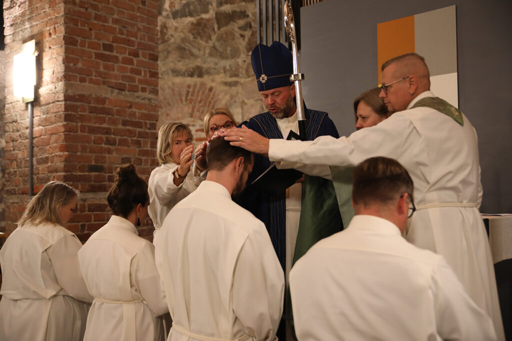Piispa ja ryhmä alpoihin pukeutuneita ihmisiä siunaa neljää alttarille polvistunutta ihmistä.